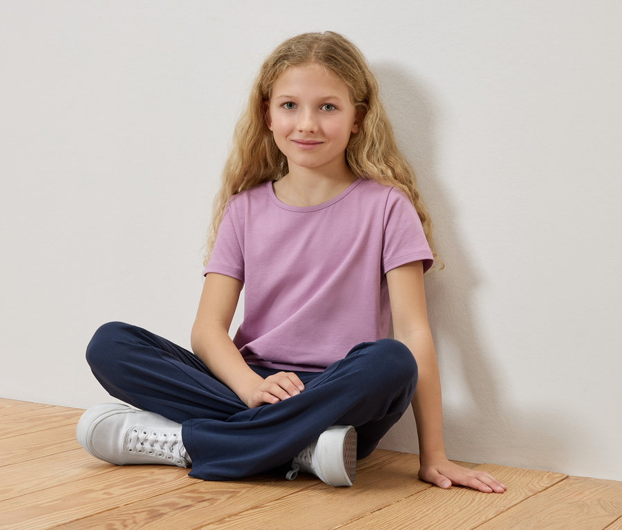 Une fille aux cheveux blonds est assise les jambes croisées sur un plancher en bois, portant un t-shirt violet et un pantalon bleu.