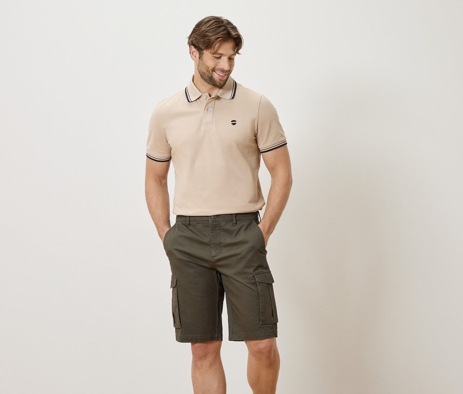 Homme portant un polo beige et un short cargo vert olive, posant les mains dans les poches sur fond blanc.