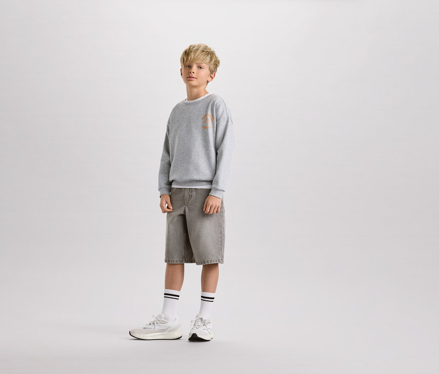 Un garçon blond se tient debout, portant un pull gris, un short et des chaussettes blanches à rayures.
