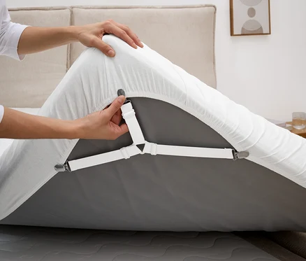 Une personne fixe un tendeur de drap blanc sur un drap-housse gris sur un matelas.