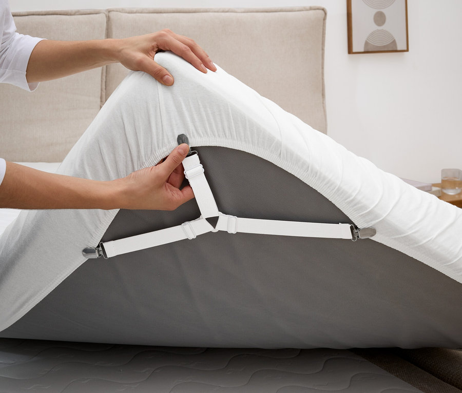 Une personne fixe un tendeur de drap blanc sur un drap-housse gris sur un matelas.