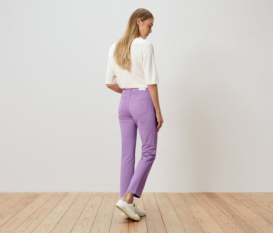 Femme vue de dos, portant un pantalon violet et un haut blanc, debout sur un plancher en bois devant un mur blanc.