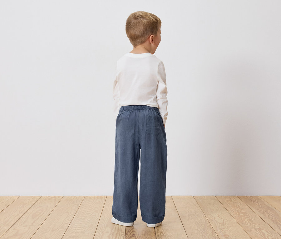 Enfant blond vu de dos, portant un t-shirt blanc à manches longues et un pantalon ample bleu.