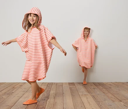 Deux enfants portant des ponchos rayés corail et blanc à capuche et des chaussures corail sur un plancher en bois devant un mur blanc.