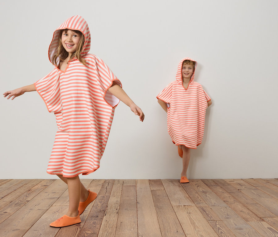 Deux enfants portant des ponchos rayés corail et blanc à capuche et des chaussures corail sur un plancher en bois devant un mur blanc.