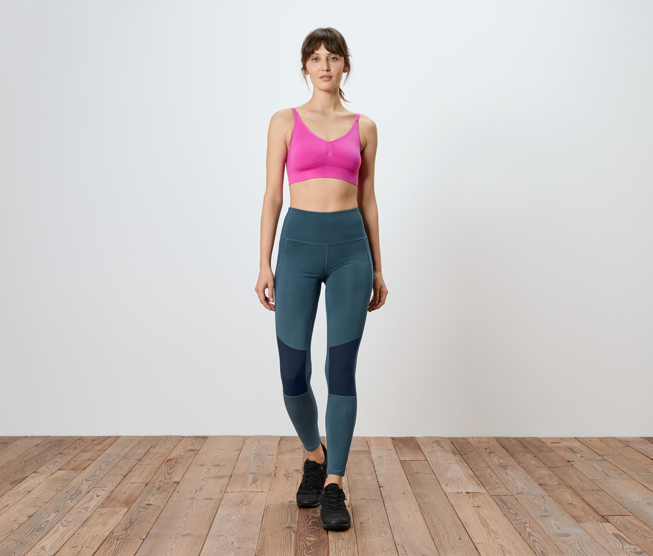 Femme marchant sur un plancher en bois, portant un soutien-gorge de sport rose et un legging bleu avec des empiècements foncés.