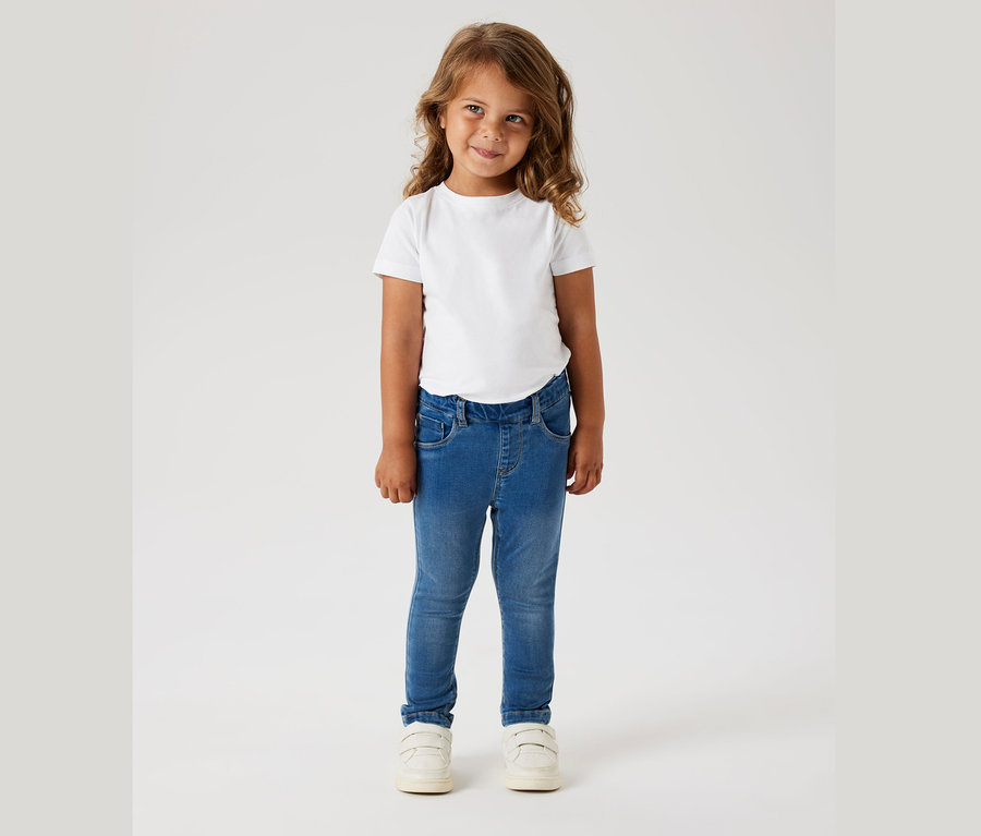Fillette aux cheveux bouclés portant un t-shirt blanc, un jean bleu et des chaussures blanches.