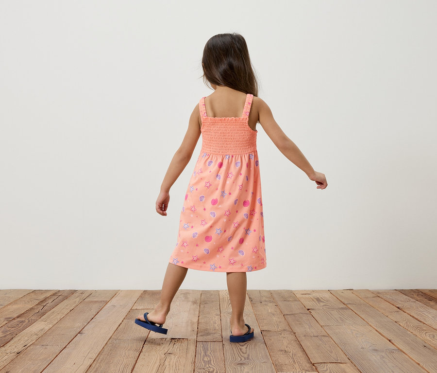 Une fille vue de dos, porte une robe d'été saumon avec des étoiles et des coquillages, avec des tongs bleues, sur un plancher en bois devant un mur blanc.