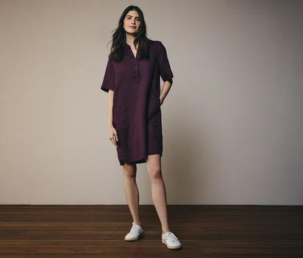 Femme portant une robe chemise bordeaux à manches courtes et des baskets blanches, debout sur un plancher en bois.