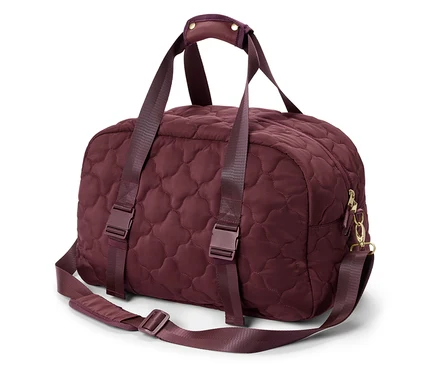 Un sac de voyage bordeaux matelassé avec des poignées et une bandoulière est posé sur un fond blanc.