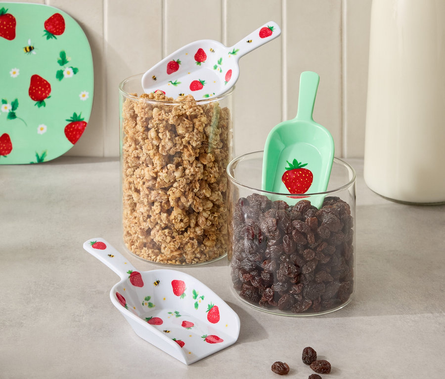 Sur la photo, deux récipients en verre remplis de granola et de raisins secs, avec une bouteille de lait entre eux. Des pelles à motif fraise sont posées sur les récipients.