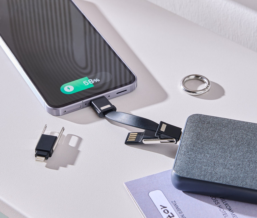 Un smartphone est en charge avec un câble et une batterie grise sur une table blanche. Une bague et une carte de chambre sont posées à côté.