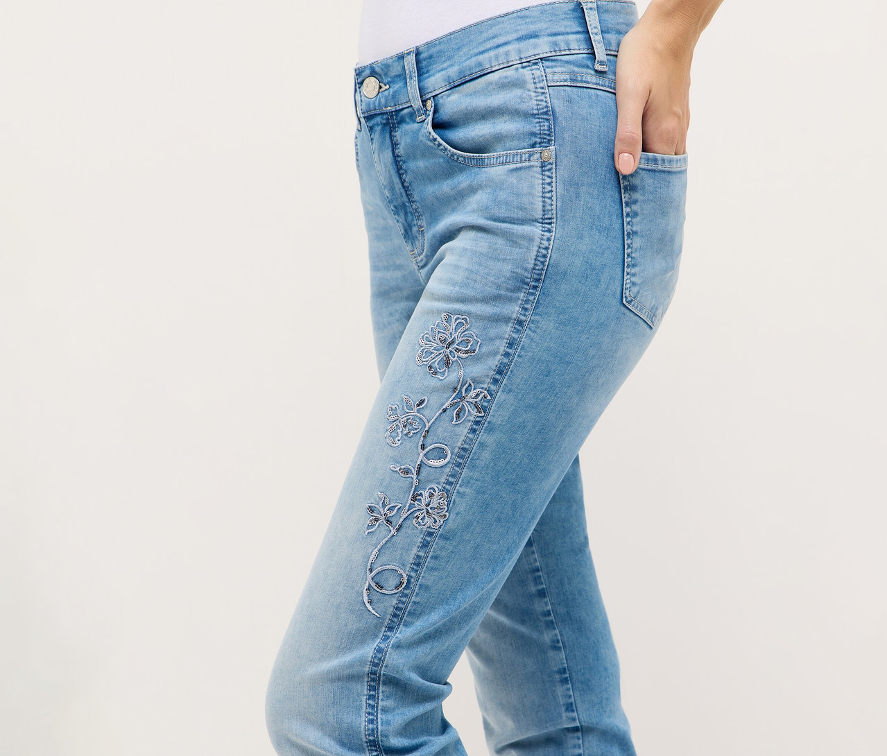 Une personne porte un jean Angels Denim « Cira Boho » avec des broderies florales et une main dans la poche.