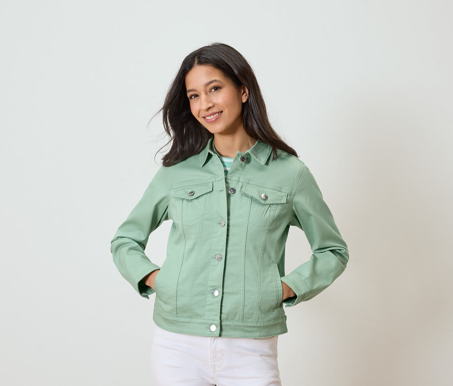 Femme posant les mains dans les poches, portant une veste en jean vert clair et un pantalon blanc.