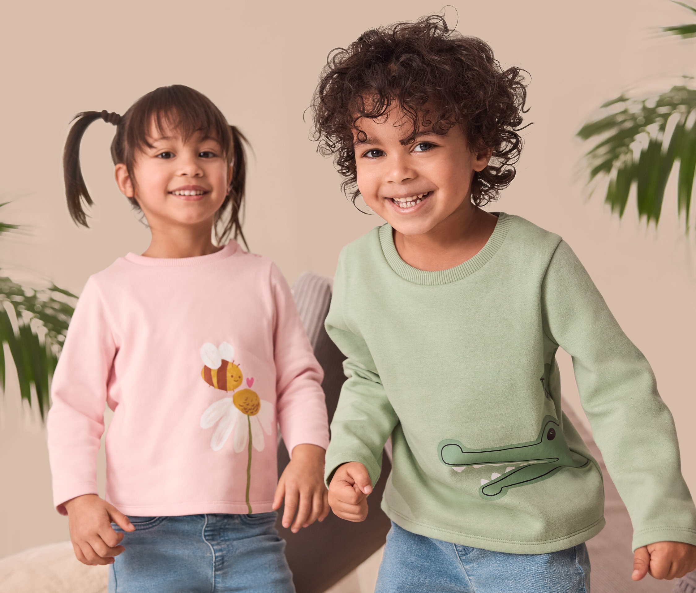 Deux enfants souriants se tiennent côte à côte. La fille à gauche porte un sweat-shirt rose pour enfants avec un imprimé de fleur et d'abeille. Le garçon à droite porte un sweat-shirt vert pour enfants avec un imprimé crocodile.