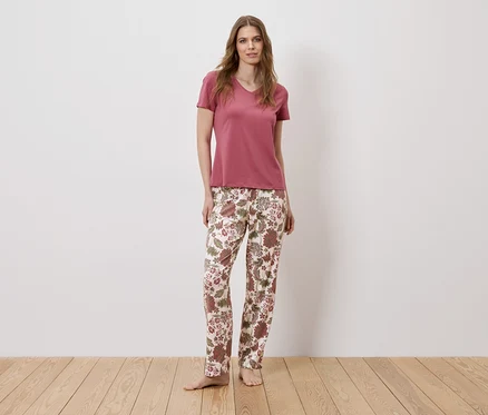 Une femme pieds nus, portant un t-shirt rose et un pantalon à fleurs, se tient devant un mur blanc.