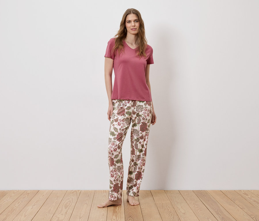 Une femme pieds nus, portant un t-shirt rose et un pantalon à fleurs, se tient devant un mur blanc.