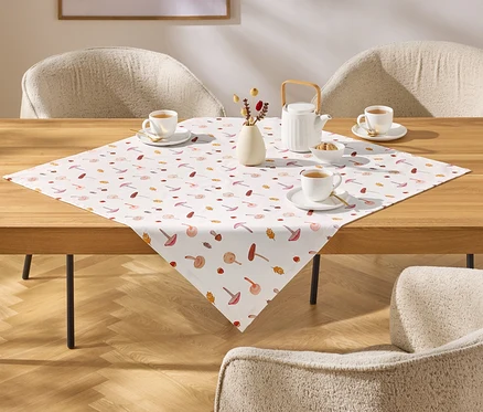 Sur une table en bois, une nappe à motifs de champignons et de feuilles, avec des tasses à thé, une théière et un vase de fleurs.
