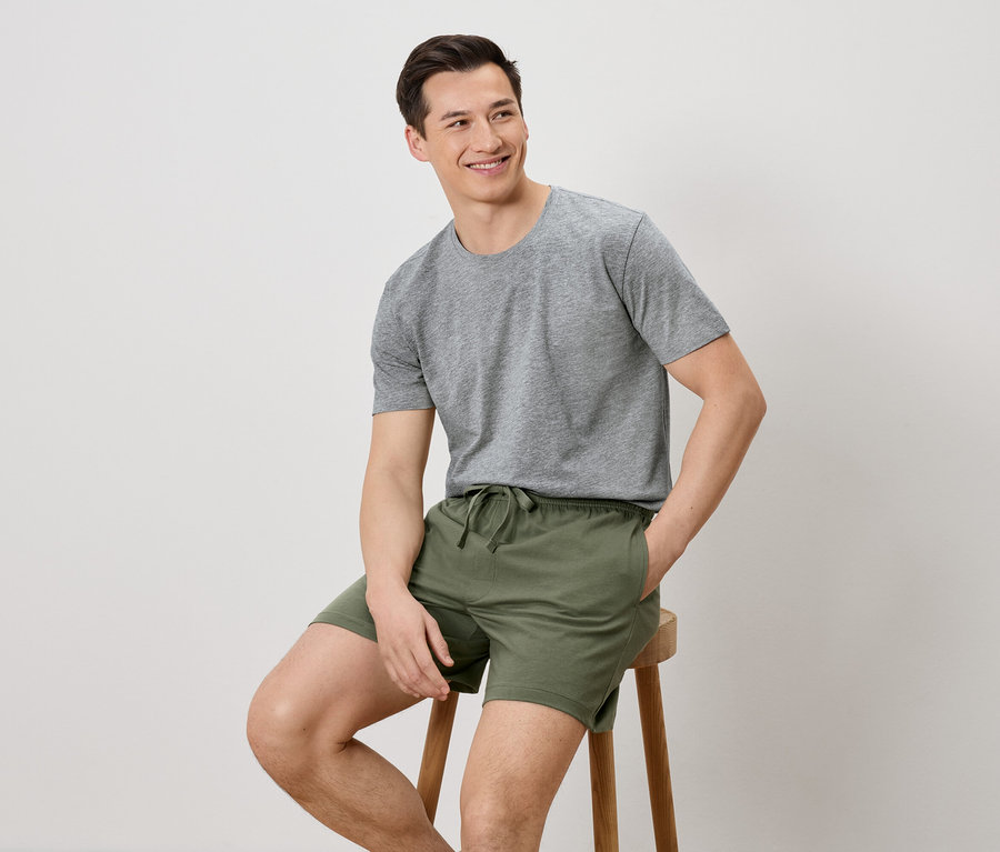 Un homme est assis sur un tabouret, portant un t-shirt gris et un short vert.
