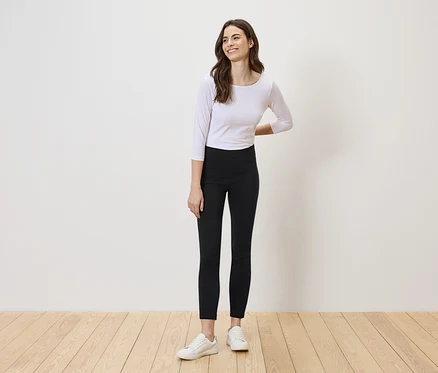 Une femme aux longs cheveux bruns se tient devant un mur blanc, portant un haut blanc, un legging noir et des baskets blanches.