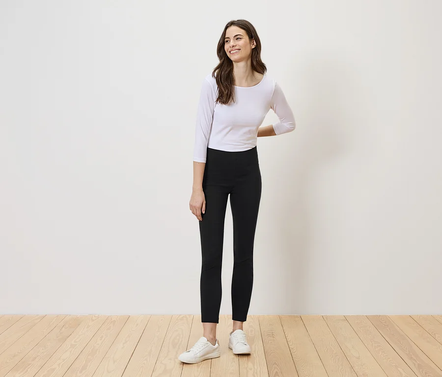 Une femme aux longs cheveux bruns se tient devant un mur blanc, portant un haut blanc, un legging noir et des baskets blanches.