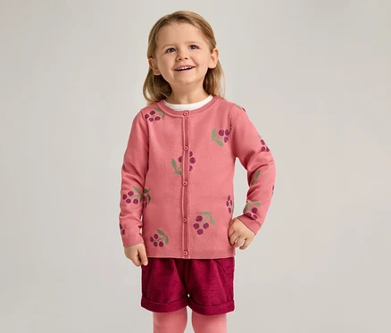 Une fillette souriante porte un cardigan rose à motif de cerises, un short bordeaux et des collants roses sur un fond clair.