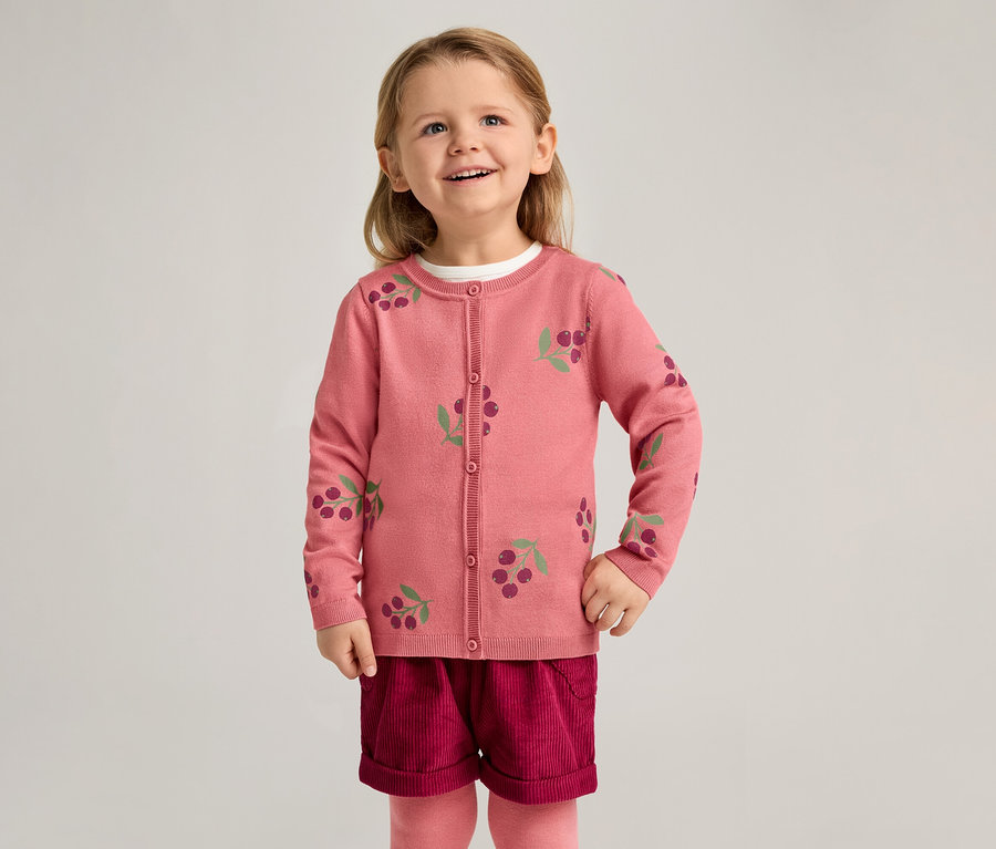 Une fillette souriante porte un cardigan rose à motif de cerises, un short bordeaux et des collants roses sur un fond clair.