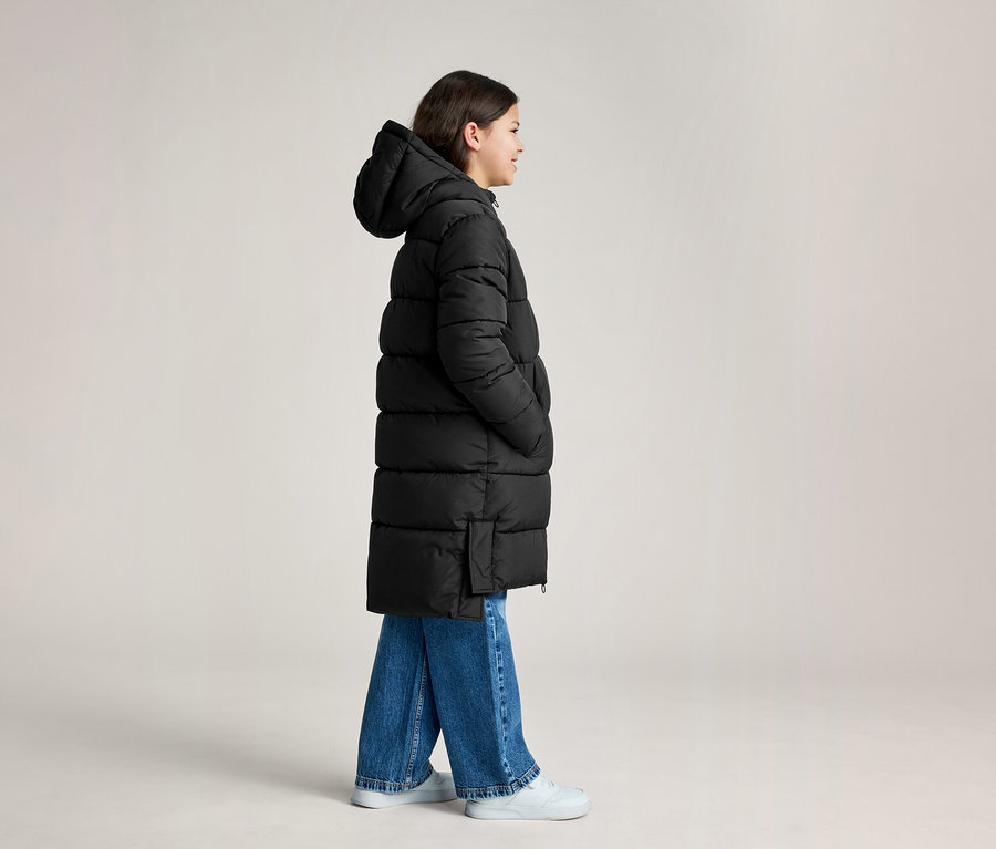 Une personne porte un long manteau doudoune noir à capuche, un jean bleu et des baskets blanches, debout de profil.