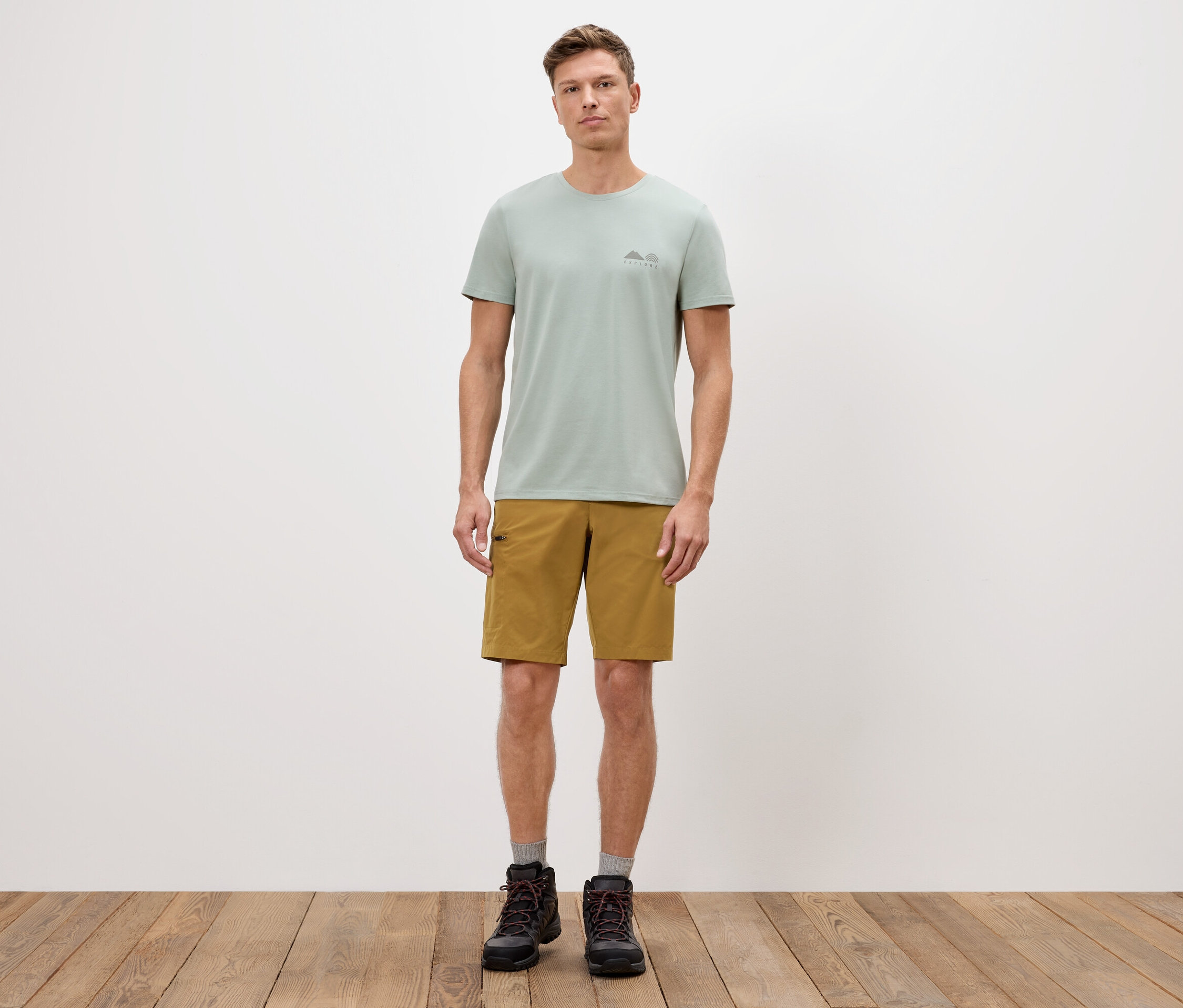 Un homme se tient devant un mur blanc, portant un t-shirt vert, un short et des bottes.