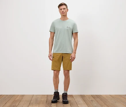 Un homme se tient devant un mur blanc, portant un t-shirt vert, un short et des bottes.
