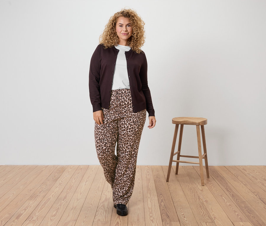 Une femme aux cheveux bouclés porte un cardigan marron, un t-shirt blanc et un pantalon à imprimé léopard.