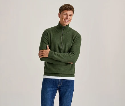 Homme souriant portant un pull vert à demi-zip sur un t-shirt blanc et un jean bleu, les bras croisés.