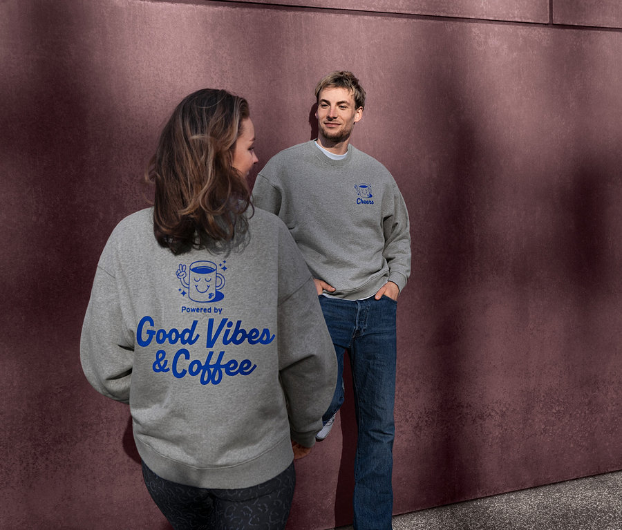 Femme et homme portant des sweat-shirts unisexe Coffee Crew gris. Le dos du sweat-shirt de la femme affiche un motif bleu "Powered by Good Vibes & Coffee". L'homme regarde la femme, son sweat-shirt a un motif bleu "Cheers".