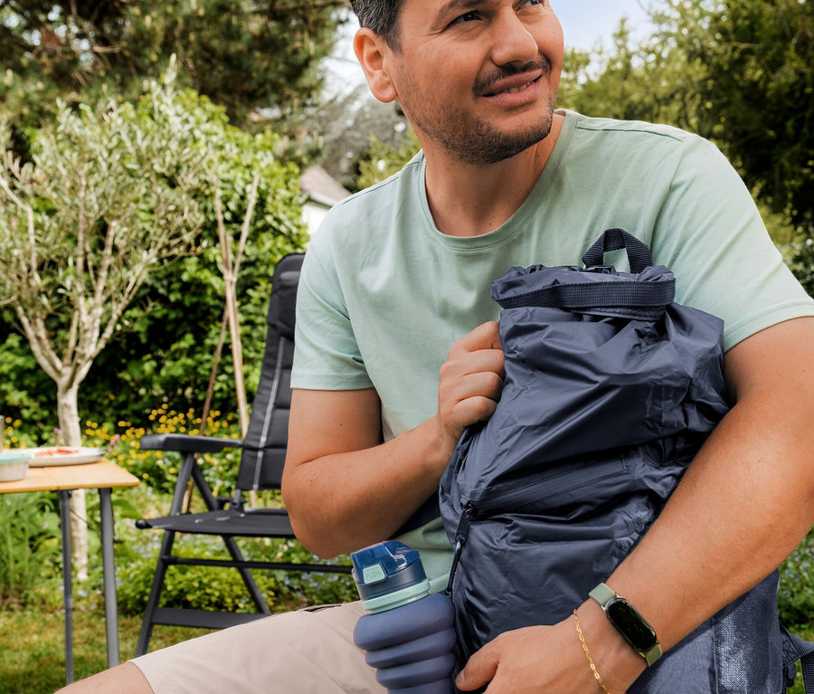 Un homme en T-shirt d’extérieur vert menthe et short beige est assis dans un jardin, tenant un sac à dos étanche bleu foncé et une gourde pliable bleue.