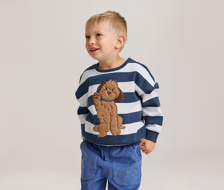 Un jeune garçon sourit et regarde vers le haut, portant un sweat-shirt rayé blanc et bleu marine avec une application de chien marron et un pantalon en velours côtelé bleu.