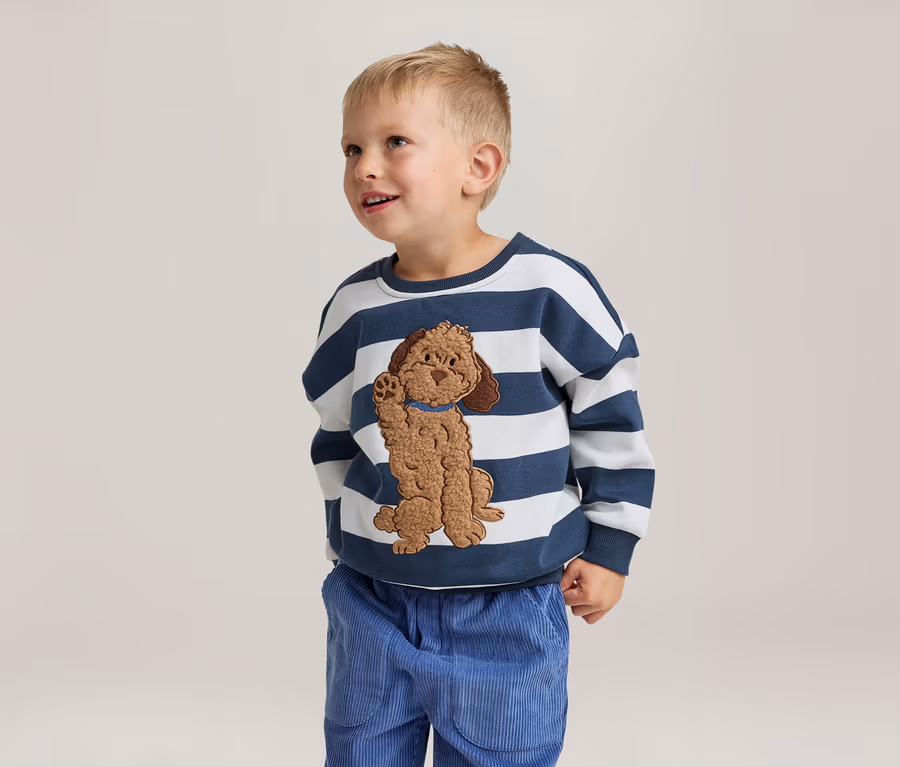 Un jeune garçon sourit et regarde vers le haut, portant un sweat-shirt rayé blanc et bleu marine avec une application de chien marron et un pantalon en velours côtelé bleu.