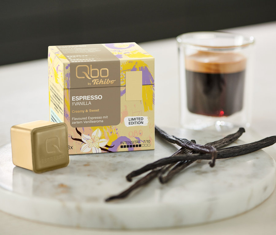 Une boîte de Qbo Flavoured Espresso Vanilla – 8 capsules, une capsule, un expresso et des gousses de vanille sur un plateau en marbre à côté d'un verre.