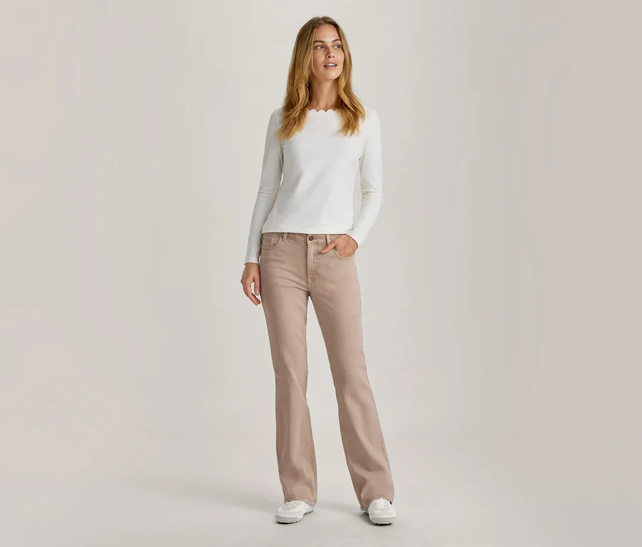 Femme debout portant un haut blanc à manches longues avec un col festonné et un jean évasé beige, une main dans la poche.