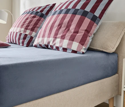 Un lit avec une tête de lit et un cadre rembourrés beiges, recouverts d'un drap-housse bleu-gris. Deux grands oreillers et un petit oreiller à carreaux rouges, blancs et bleus sont posés sur le lit. Une lampe blanche et une table de chevet sont visibles en arrière-plan.