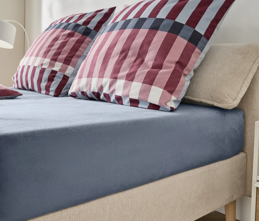 Un lit avec une tête de lit et un cadre rembourrés beiges, recouverts d'un drap-housse bleu-gris. Deux grands oreillers et un petit oreiller à carreaux rouges, blancs et bleus sont posés sur le lit. Une lampe blanche et une table de chevet sont visibles en arrière-plan.