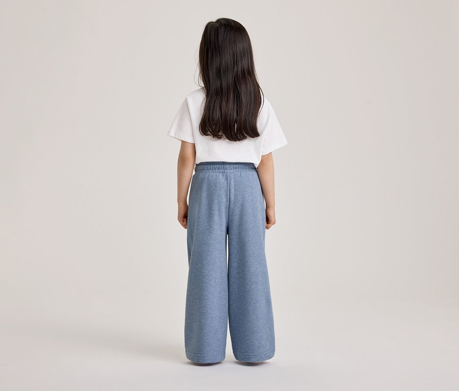 Enfant aux longs cheveux foncés, vu de dos, portant un t-shirt blanc et un pantalon de jogging large bleu.