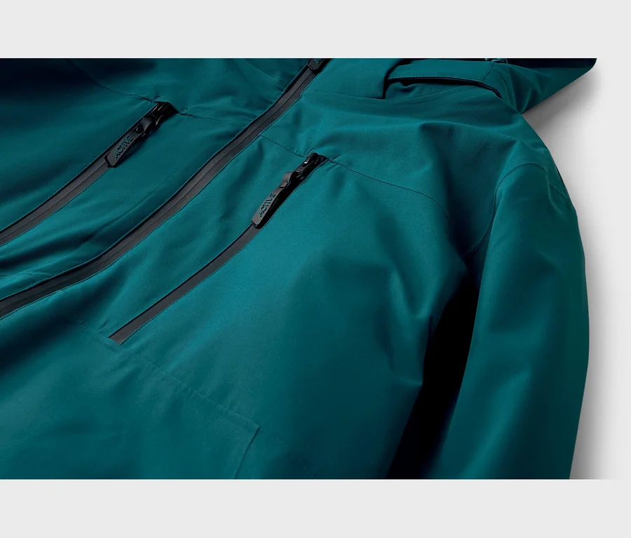 Gros plan d'une veste sarcelle avec des fermetures éclair noires et l'inscription 'ACTIVE' sur les tirettes. La veste a plusieurs poches zippées et une capuche visible.