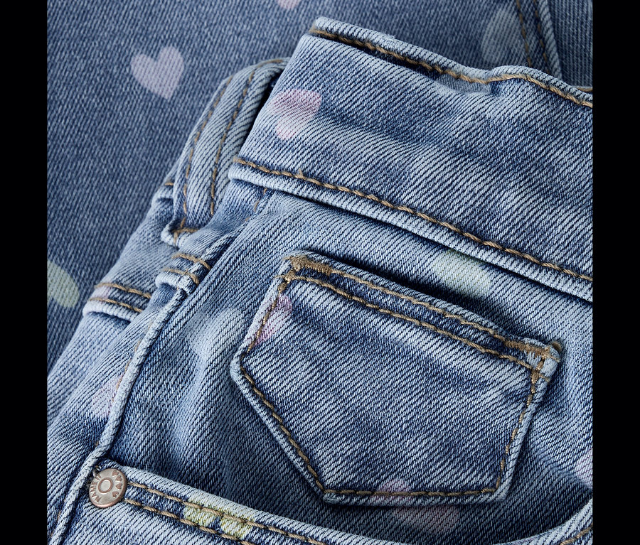 Gros plan d'un jean en denim bleu clair avec des motifs de cœurs roses, vert clair et violet clair dispersés. Une poche arrière et la ceinture avec des surpiqûres beiges sont visibles.
