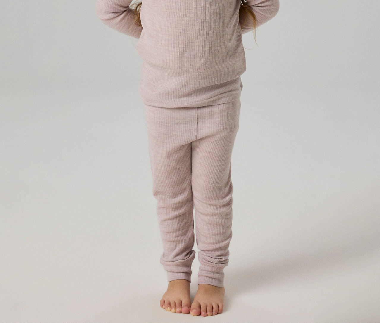 Enfant debout pieds nus, portant un haut côtelé rose clair à manches longues et un legging côtelé assorti.