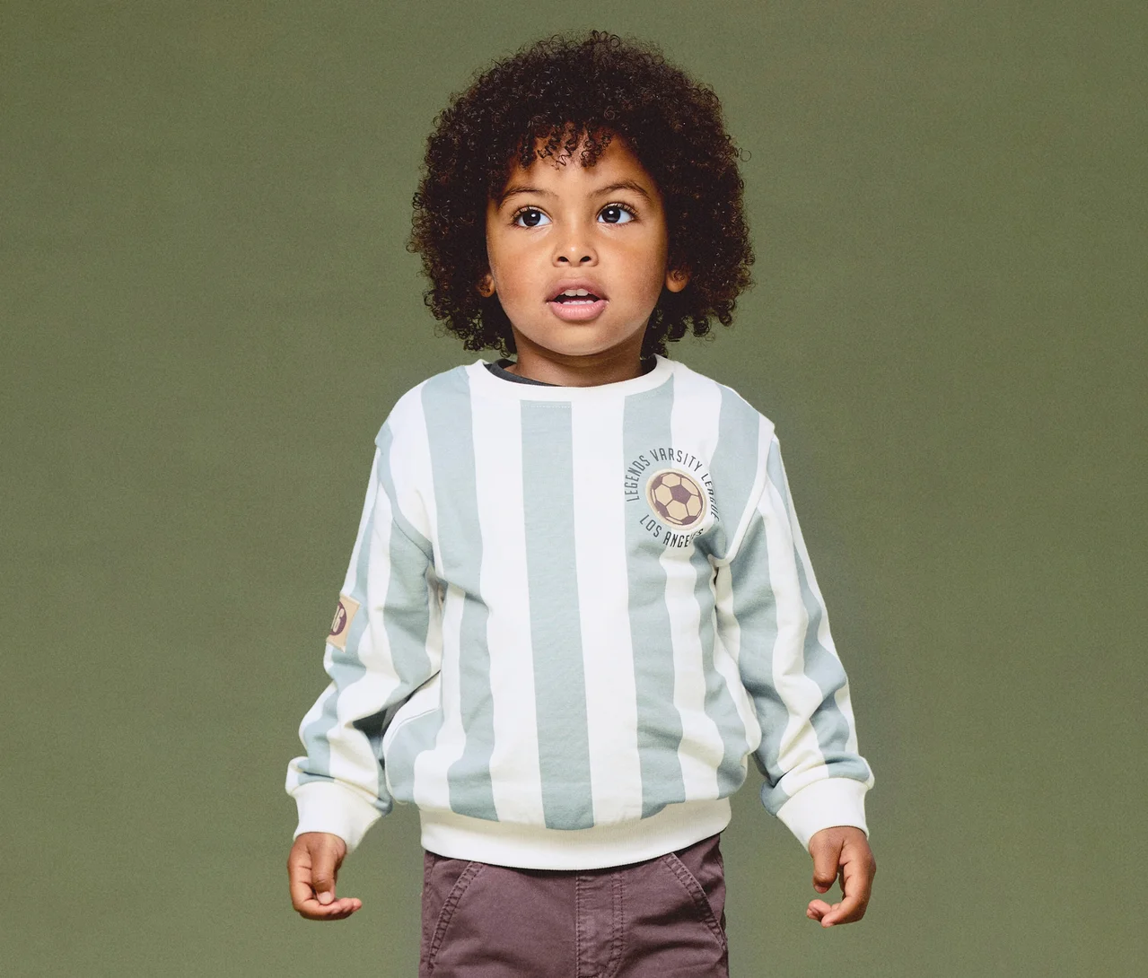 Un jeune garçon aux cheveux bouclés foncés porte un sweat-shirt rayé vert clair et blanc cassé avec un emblème de ballon de football et un pantalon marron, regardant vers le haut sur un fond vert.
