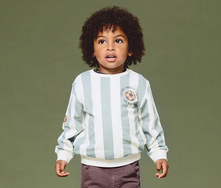 Un jeune garçon aux cheveux bouclés foncés porte un sweat-shirt rayé vert clair et blanc cassé avec un emblème de ballon de football et un pantalon marron, regardant vers le haut sur un fond vert.