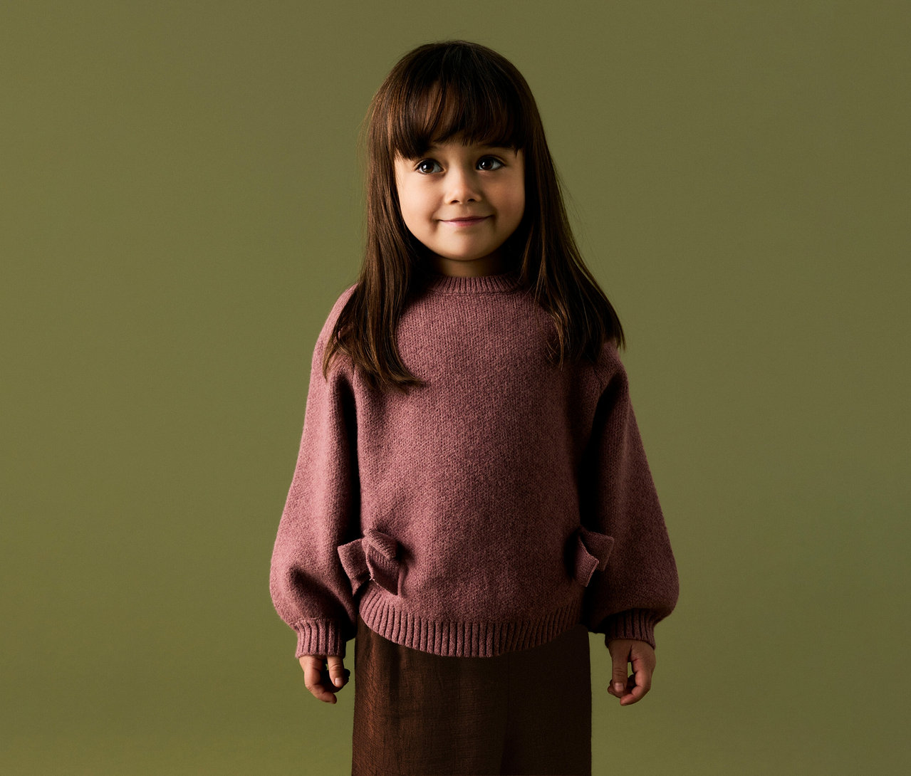 Jeune enfant aux longs cheveux foncés et frange, souriant, portant un pull tricoté rose poudré à manches bouffantes et détails nœuds, et un pantalon marron foncé, sur fond vert olive.