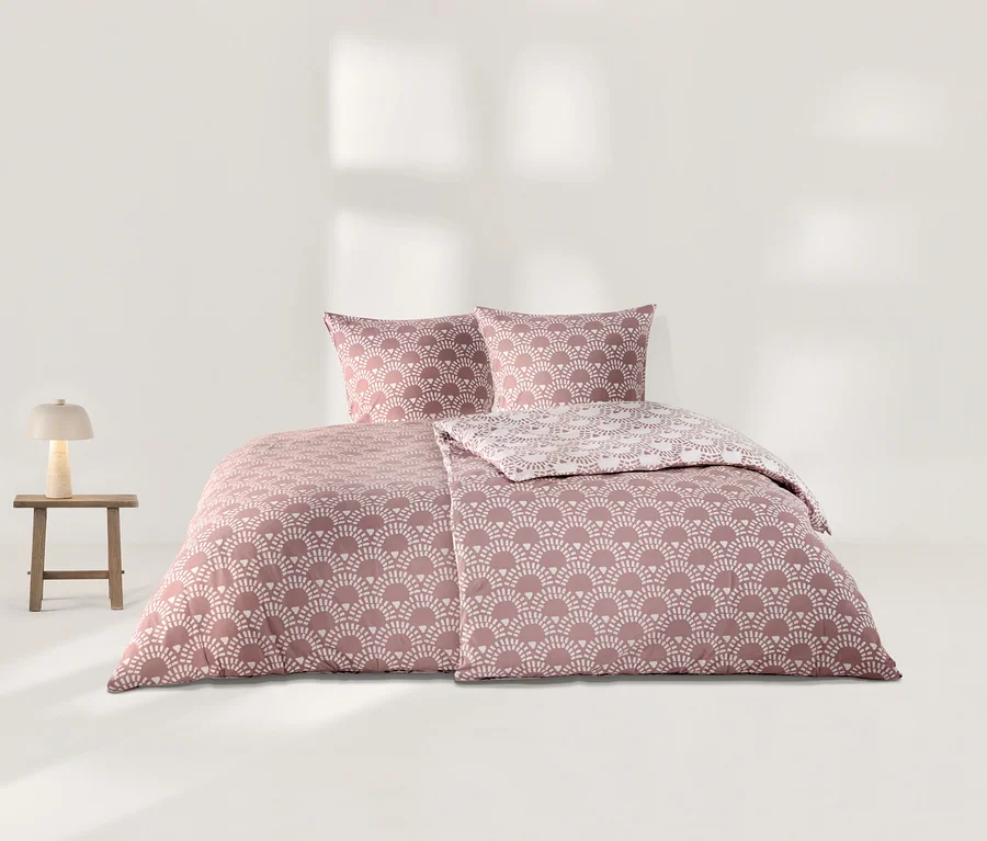 Parure de lit en jersey rose à motif d'éventails blancs, comprenant deux oreillers et une housse de couette dont un côté présente un motif plus clair. À gauche, un tabouret en bois avec une lampe beige.