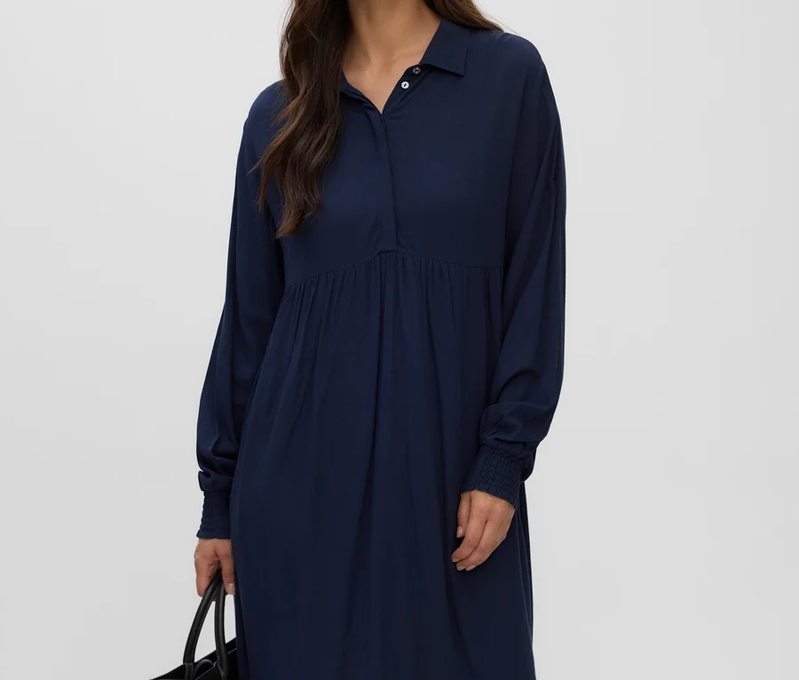 Femme aux longs cheveux foncés, portant une robe MAMALICIOUS « ESRA LIA » bleu marine avec col, boutons et taille froncée, tenant un sac noir.