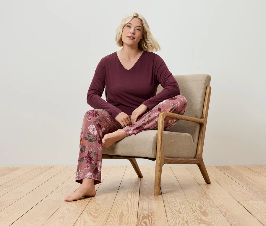 Une femme blonde est assise dans un fauteuil et porte un T-shirt bordeaux à manches longues et un pantalon de pyjama à motifs floraux.